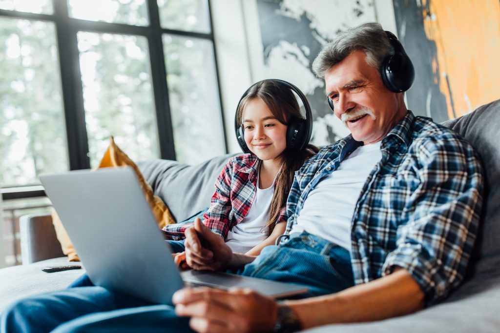 Foto de abuelo y nieta escuchando música de un ordenador con auriculares, sentados en el sofá.